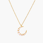 Gold crescent moon pendant with graduated white pearls on delicate chain against cream background