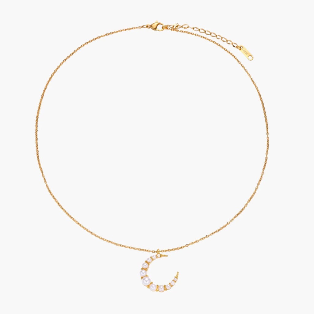 Gold crescent moon pendant with graduated white pearls on delicate chain against cream background