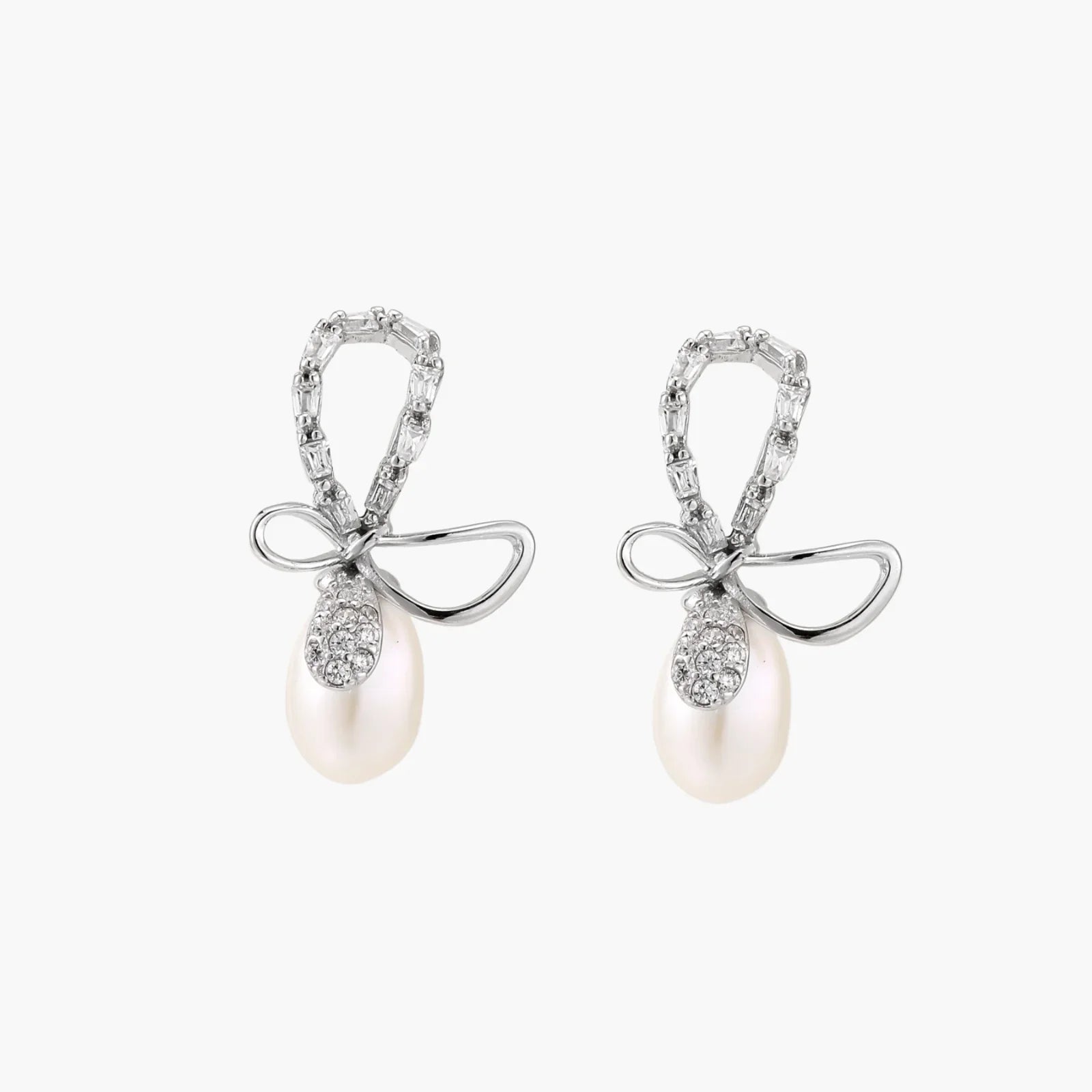 Silver bow-shaped stud earrings with baguette crystals and dangling white freshwater pearls on cream background